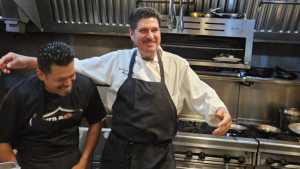 A vibrant dog park bathed in sunlight features abundant greenery with mature trees, offering ample shade for leisurely outdoor activities. In the distance is a well-equipped playground that presents an additional activity area. An inviting picnic table spreads out nearby, ideal for enjoying meals al fresco or taking relaxing breaks from play. A neatly-placed trash bin aids in maintaining the park's cleanliness for everyone's enjoyment. Dominating the scene is a generously wide dirt path gracefully curving through the expanse of grass, promising exploratory walks and lively fetch games.